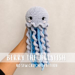 May include: A crochet jellyfish toy with a light grey rounded head, black safety eyes, and pink embroidered cheeks. It features long, curly tentacles in alternating shades of light grey and bright blue. Text reads: BERRY THE JELLYFISH NO SEW CROCHET PATTERN.