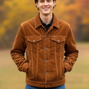 Könnte beinhalten: Braune Cordjacke mit zwei Brusttaschen und zwei Seitentaschen. Die Jacke hat eine Knopfleiste und ein kleines rotes Etikett auf der Brust. Die Person trägt ein schwarzes Hemd unter der Jacke.