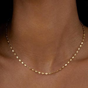 Könnte beinhalten: Eine zarte goldene Halskette mit einem sich wiederholenden rautenförmigen Design. Die Halskette wird um den Hals getragen und liegt auf dem Schlüsselbein. Der Hintergrund ist dunkel und hebt den Schmuck hervor.