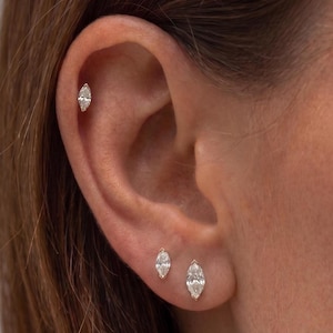 May include: Three marquise-cut clear gemstone stud earrings set in gold metal. One earring is positioned in the upper cartilage, while two earrings of varying sizes are placed in the earlobe, creating a coordinated set of jewellery pieces.