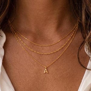 Könnte beinhalten: Ein geschichtetes Goldhalsketten-Set bestehend aus drei zarten Ketten mit unterschiedlichen Texturen. Die längste Kette verfügt über einen kleinen, mittig platzierten goldenen Anhänger in Form des Großbuchstabens A. Die Ketten sind dünn und poliert.