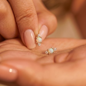 May include: A pair of gold stud earrings, each featuring a central round opal cabochon flanked by two small, clear, round-cut accent stones. The design is symmetrical, with the opal positioned between the two smaller stones in a linear arrangement.