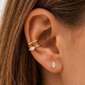 Könnte beinhalten: Ein goldener Ohrring-Cuff mit zwei parallelen Bändern, wobei das untere Band mit einem klaren Stein im Marquise-Schliff verziert ist, kombiniert mit einem passenden goldenen Ohrstecker, der einen einzelnen klaren Marquise-Stein enthält.