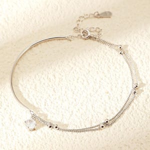 Puede incluir: Una tobillera plateada con un diseño delicado. Presenta una barra curva, un pequeño colgante de piedra preciosa transparente y dos hilos de cadena. Un hilo tiene pequeñas cuentas redondas. La cadena ajustable permite un ajuste personalizado.