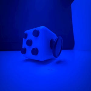 May include: A white fidget cube with black tactile features, including four small circular buttons on the front face and a larger circular dial on the side, all illuminated under a strong blue light source in a dark environment.