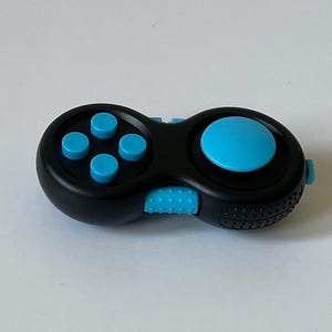 Puede incluir: Un controlador fidget negro con detalles azules. El controlador tiene un botón circular y cuatro botones más pequeños en un lado, y un botón circular más grande en el otro. El centro presenta un interruptor azul texturizado.