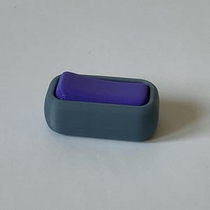May include: A small, rectangular, grey container with rounded edges holds a stack of purple, folded material. The container is a solid, matte colour, and the purple material contrasts with the grey. The item appears to be a desk organiser or storage solution.