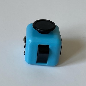 May include: A light blue fidget cube with black buttons and a black joystick. The cube is designed to help with focus and stress relief. The cube has several different sides with various tactile features.