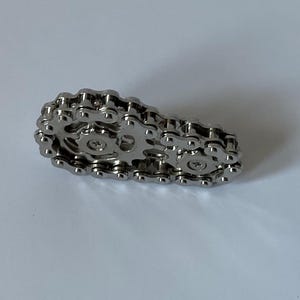 Puede incluir: Un diseño de eslabón de cadena de bicicleta de color plateado. La cadena de metal está dispuesta en forma rectangular, con engranajes y eslabones de conexión visibles. El objeto está sobre una superficie blanca.