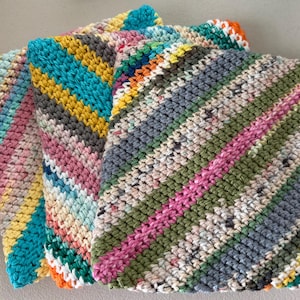 Puede incluir: Tres paños cuadrados de ganchillo coloridos. Cada paño presenta un patrón de rayas diagonales en varios colores, incluyendo rosa, verde, azul, amarillo y gris. La superficie texturizada y los tonos vibrantes sugieren un artículo hecho a mano, posiblemente para la cocina o el hogar.