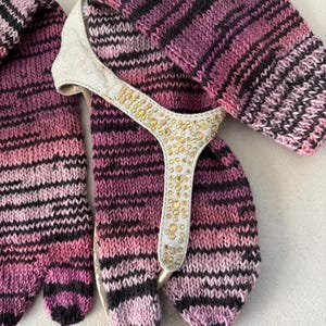 May include: A pair of knitted gloves and a matching sandal. The gloves and sandal are striped with shades of pink, black, and purple. The sandal has a white strap adorned with gold-colored embellishments.