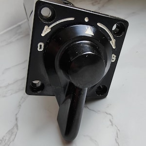 May include: A black control switch with a square base and a rounded knob. The base has white markings and screw holes. The switch has a lever and is likely used for electrical or mechanical control.