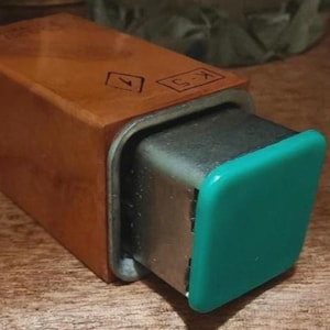 May include: A rectangular wooden box with a metal insert and a teal-colored cap. The box has a light brown finish and is marked with symbols and the text "K-5". The object is on a wooden surface.