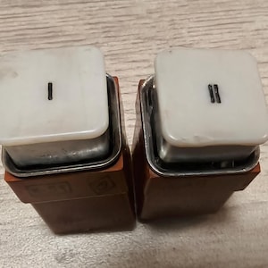 May include: Two vintage electrical components. Each has a white, square top with a black Roman numeral "I" or "II". The tops sit on a metal frame and a brown base. The items are displayed on a light wood surface.