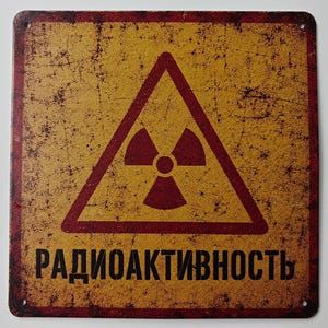 Peut inclure: Une plaque métallique carrée avec une finition vieillie et usée jaune et rouge foncé. Elle présente un symbole de danger radioactif rouge foncé au centre dans un triangle, avec le texte cyrillique "РАДИОАКТИВНОСТЬ" en lettres capitales noires.