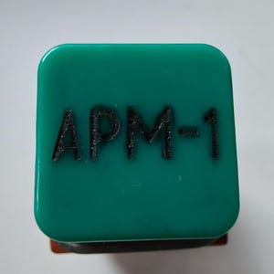 Pode incluir: Um objeto quadrado de cor verde-azulado com cantos arredondados, apresentando o texto preto em relevo "APM-1" centralizado na sua superfície superior. O objeto tem um acabamento liso e brilhante e uma estrutura sólida com uma base escura.