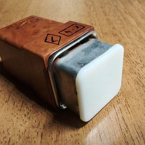 May include: A rectangular, brown wooden object with a white square button on one end. The object has a metal frame and is resting on a wooden surface. The text "K-4" is visible on the top.