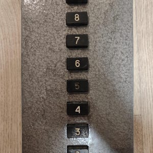 Könnte beinhalten: Ein rechteckiges Aufzugsbedienfeld mit einer texturierten grauen Metalloberfläche. Das Panel verfügt über eine vertikale Reihe von neun schwarzen quadratischen Tasten, die jeweils mit weißen Ziffern von 1 bis 9 in aufsteigender Reihenfolge beschriftet sind.