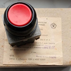 May include: A red button with a black bezel sits atop a gray base, resting on a cardboard box. The button has the text "KEO" on its side. A paper label with Cyrillic text is visible.