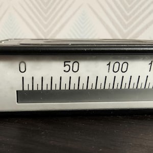 Könnte beinhalten: Ein schwarzes, rechteckiges analoges Voltmeter mit weißer Frontplatte und einer horizontalen Skala von 0 bis 200. Das Gerät zeigt auf der linken Seite einen großen Buchstaben V für Spannungsmessung und verfügt über eine lineare Anzeige.