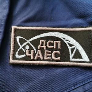 Pode incluir: Um patch retangular com o texto "ДСП ЧАЕС" em letras castanhas claras. O patch apresenta um símbolo de átomo branco e uma estrutura estilizada sobre um fundo preto, emoldurado por uma borda castanha clara. O patch é costurado em um tecido azul.