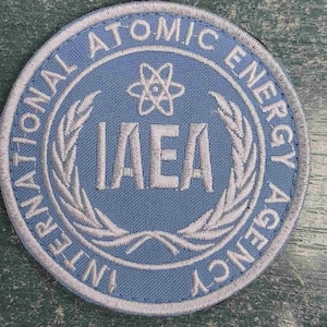 May include: A circular patch with a light blue background and white embroidered details. The text "INTERNATIONAL ATOMIC ENERGY AGENCY" surrounds the center, which features the letters "IAEA" and a stylized atom symbol.