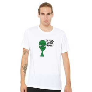Puede incluir: Camiseta blanca con un alienígena verde con expresión de enfado y el texto "OH FUCK. WRONG PLANET". La camiseta está hecha de un material suave, ideal para uso casual.