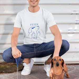 Peut inclure: T-shirt blanc avec un dessin bleu d'un chien portant des lunettes de soleil et le texte "FETCH THIS". Un chien teckel marron avec un harnais marron est en laisse. La personne porte un jean bleu et des baskets blanches.