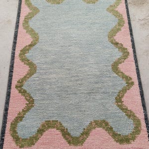 May include: A rectangular area rug featuring a central light blue section with a wavy, scalloped olive green border. The surrounding field is light pink, enclosed by a dark gray border with white fringe tassels along the top and bottom edges.