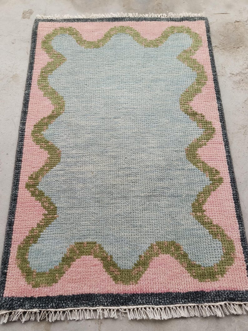 May include: A rectangular area rug featuring a central light blue section with a wavy, scalloped olive green border. The surrounding field is light pink, enclosed by a dark gray border with white fringe tassels along the top and bottom edges.