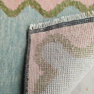May include: A woven area rug featuring a light blue base with a repeating wavy pattern in soft pink and olive green. The rug has a dark gray border and a white fringed edge where the corner is folded over to reveal the textured underside.
