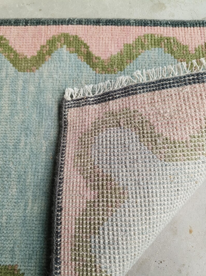 May include: A woven area rug featuring a light blue base with a repeating wavy pattern in soft pink and olive green. The rug has a dark gray border and a white fringed edge where the corner is folded over to reveal the textured underside.