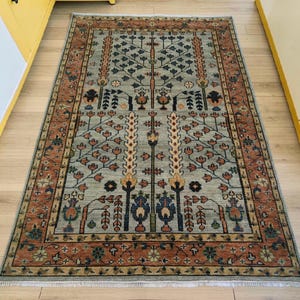 May include: A rectangular area rug featuring a light sage green field with a central tree of life motif in shades of blue, orange, and cream. The rug is framed by a wide, rust-coloured border decorated with repeating floral and vine patterns.