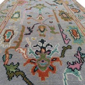 May include: A rectangular, gray rug with an intricate floral pattern. The rug features a central design with orange, green, and black elements, surrounded by smaller floral motifs in pink, teal, and other colors. The border is a warm, muted orange.