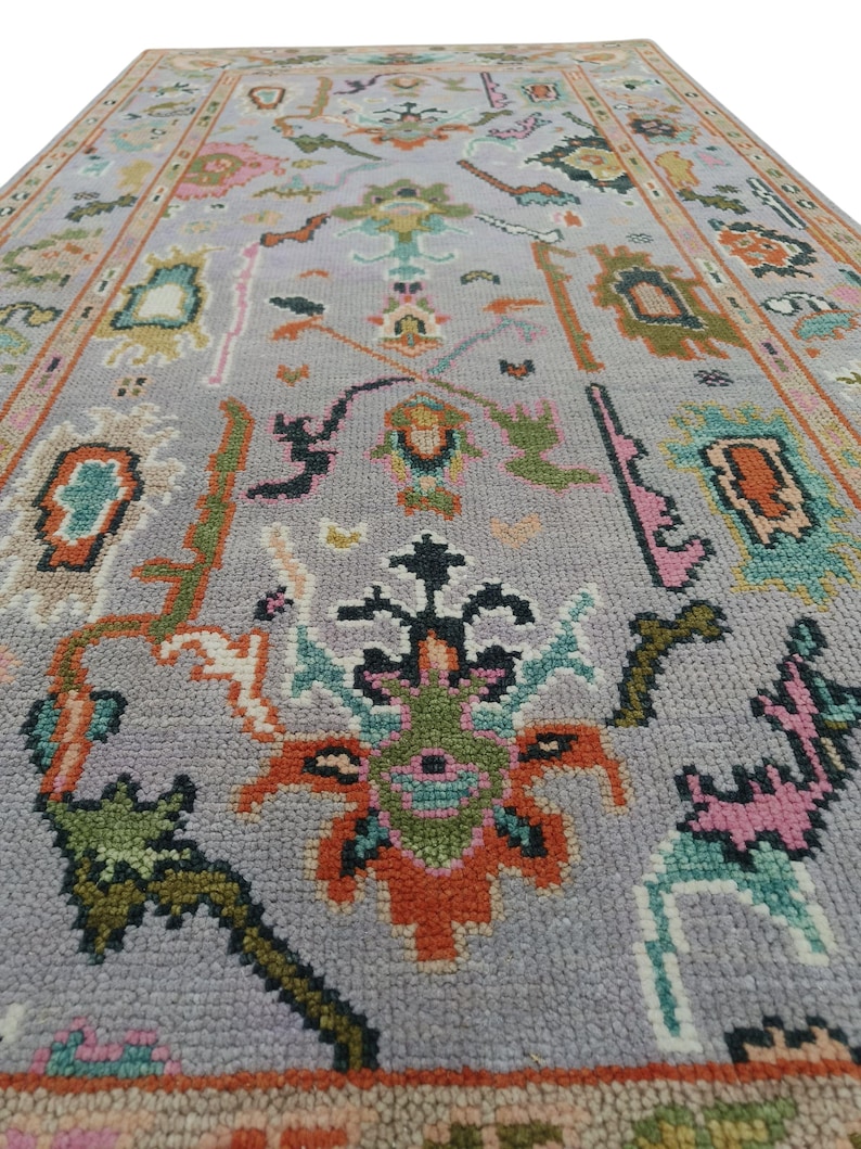 May include: A rectangular, gray rug with an intricate floral pattern. The rug features a central design with orange, green, and black elements, surrounded by smaller floral motifs in pink, teal, and other colors. The border is a warm, muted orange.