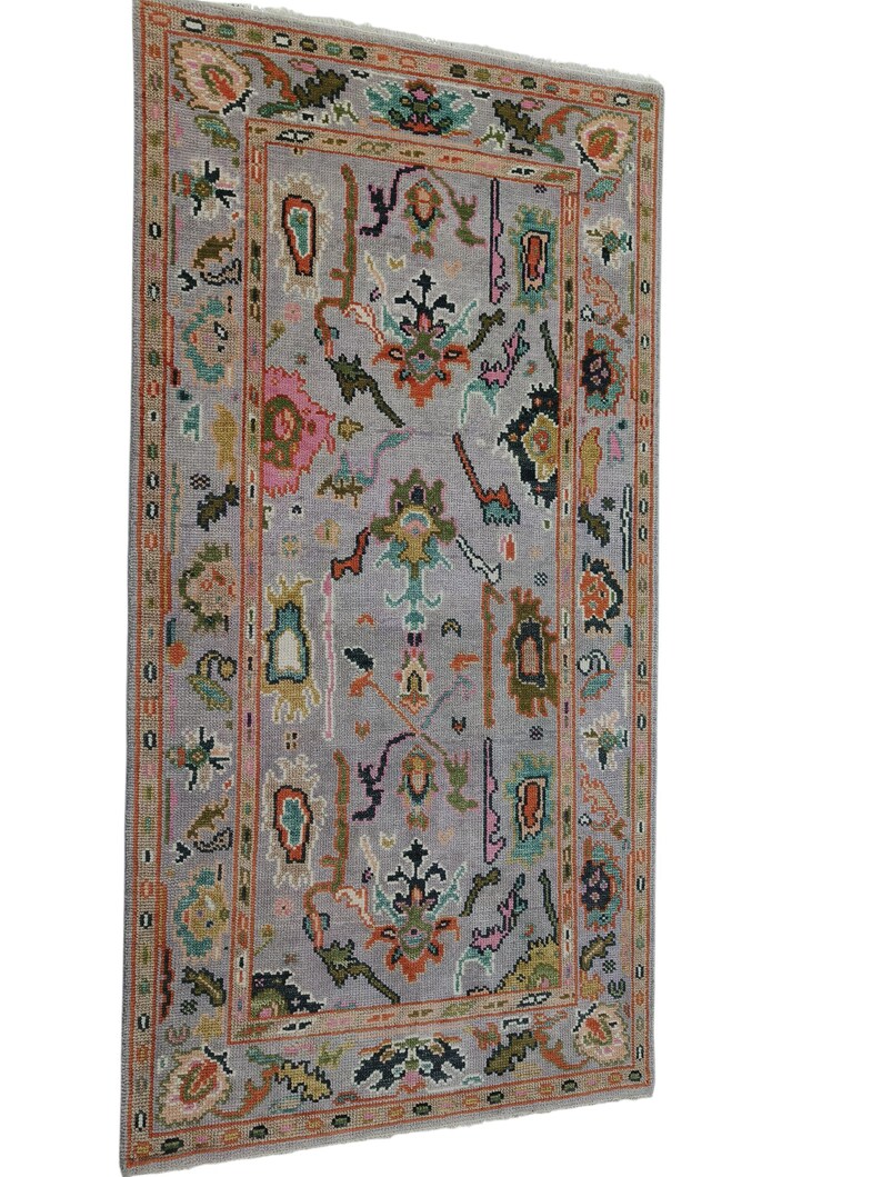 May include: A rectangular, gray rug with an intricate, colorful pattern. The design features floral and geometric shapes in shades of pink, green, orange, and blue. The rug has a detailed border and is likely made of woven material.