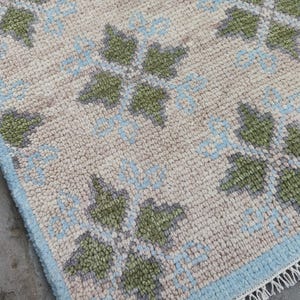 May include: Close-up of a beige rug with a repeating pattern of green and light blue floral designs. The rug has a light blue border and fringe along one edge. The rug is made of a textured material.