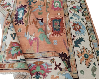 May include: A peach-toned area rug with a detailed, colourful pattern. The rug features a central field with various shapes and designs in shades of green, blue, and red. A cream-coloured border frames the rug, with additional patterns and colours.