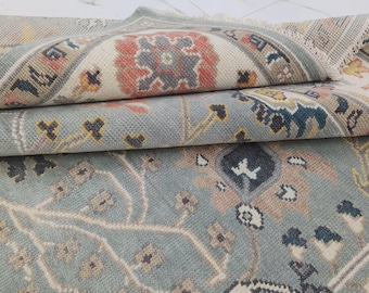 May include: A folded area rug featuring a light blue base with intricate floral and geometric patterns in shades of cream, terracotta, dark gray, and yellow. The rug has a visible woven texture and short fringe along the folded edge.