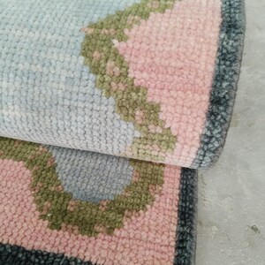 May include: A hand-knotted wool rug featuring a light blue center with a scalloped border in olive green and soft pink. The rug has a dark charcoal gray outer edge and a short white fringe along the bottom, showcasing a dense, textured pile.