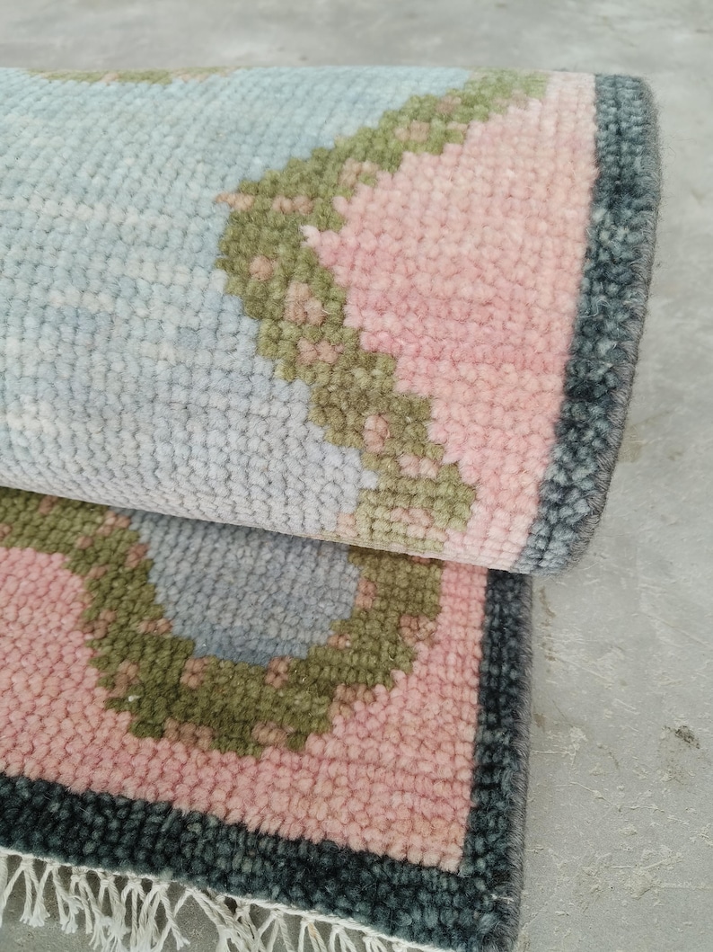 May include: A hand-knotted wool rug featuring a light blue center with a scalloped border in olive green and soft pink. The rug has a dark charcoal gray outer edge and a short white fringe along the bottom, showcasing a dense, textured pile.