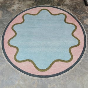 May include: A circular area rug featuring a central light blue scalloped shape outlined in olive green. The rug has a wide pink border surrounding the blue center and is finished with a thin dark gray outer edge and a light-colored fringe trim.