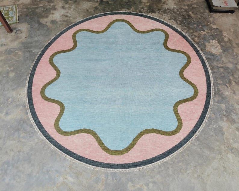 May include: A circular area rug featuring a central light blue scalloped shape outlined in olive green. The rug has a wide pink border surrounding the blue center and is finished with a thin dark gray outer edge and a light-colored fringe trim.