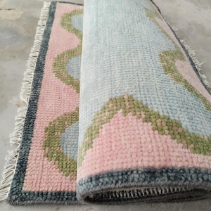May include: A rectangular area rug featuring a soft pink background with light blue scalloped shapes outlined in olive green. The rug has a dark gray border and white fringe on the short ends. The rug is partially rolled to show the textured pile.