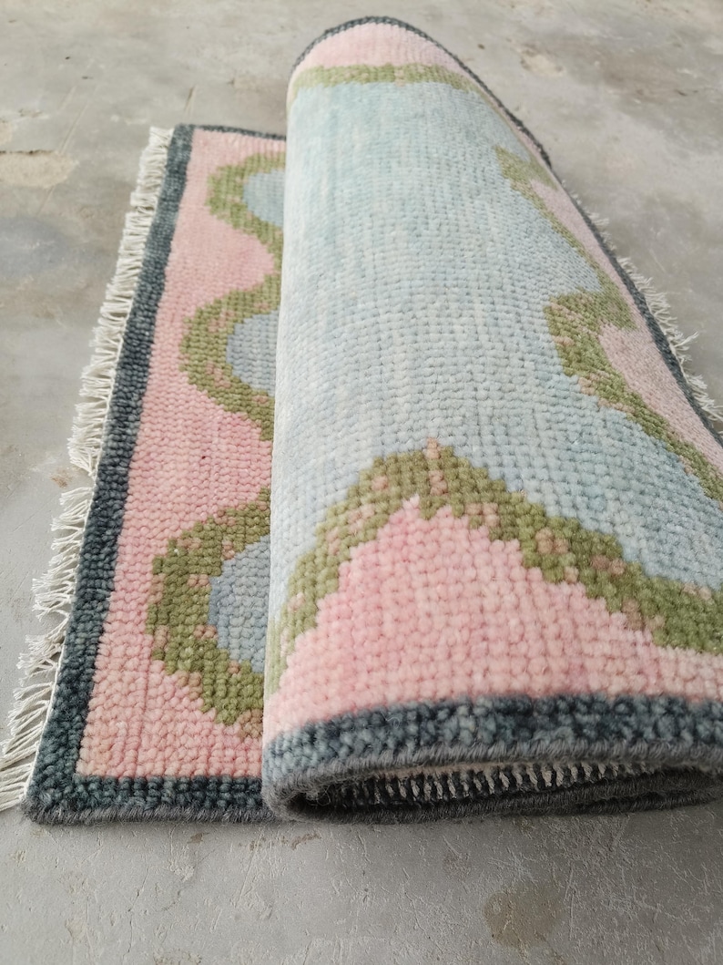 May include: A rectangular area rug featuring a soft pink background with light blue scalloped shapes outlined in olive green. The rug has a dark gray border and white fringe on the short ends. The rug is partially rolled to show the textured pile.