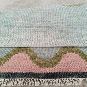 May include: A partially rolled area rug featuring a textured, tufted pile with a repeating pattern of light blue, soft pink, and olive green scalloped shapes. The rug is finished with a dark gray border and a row of white fringe along the bottom edge.