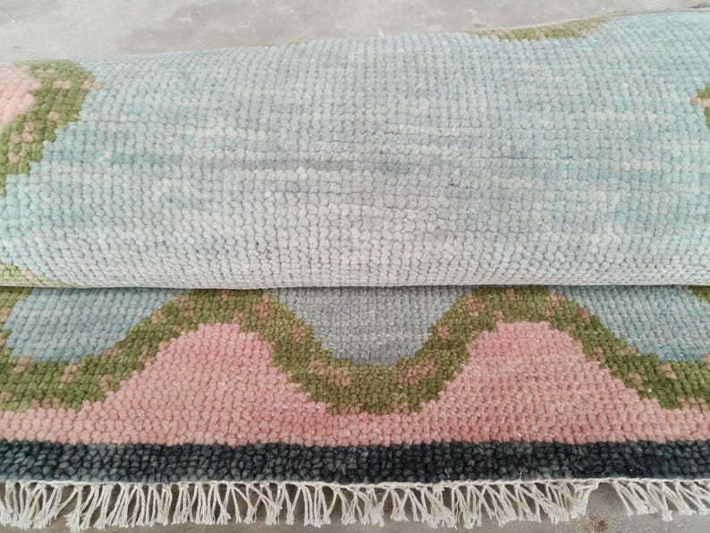 May include: A partially rolled area rug featuring a textured, tufted pile with a repeating pattern of light blue, soft pink, and olive green scalloped shapes. The rug is finished with a dark gray border and a row of white fringe along the bottom edge.