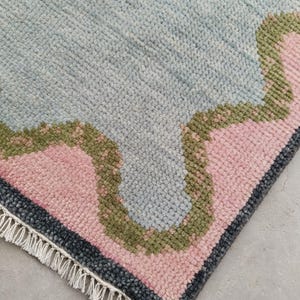 May include: A rectangular area rug featuring a thick, tufted pile texture. The design consists of a large light blue central section bordered by a wavy olive green band, followed by a soft pink area and a dark charcoal gray edge with white fringe.