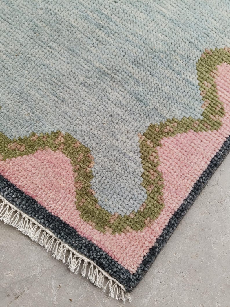 May include: A rectangular area rug featuring a thick, tufted pile texture. The design consists of a large light blue central section bordered by a wavy olive green band, followed by a soft pink area and a dark charcoal gray edge with white fringe.