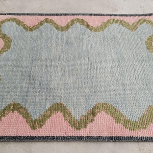 May include: A rectangular area rug featuring a central light blue field surrounded by a wavy olive green border and a pink outer frame. The rug is finished with a dark gray edge and short fringe on the left and right sides, showcasing a textured pile.